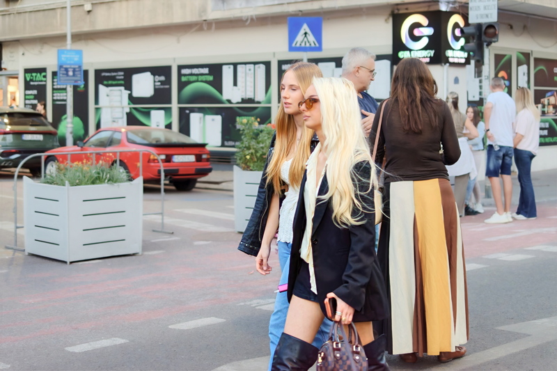 25 - Two young women walk down a sunny street together, with one wearing a black blazer and over-the-knee boots and the other in a black leather jacket and jeans
